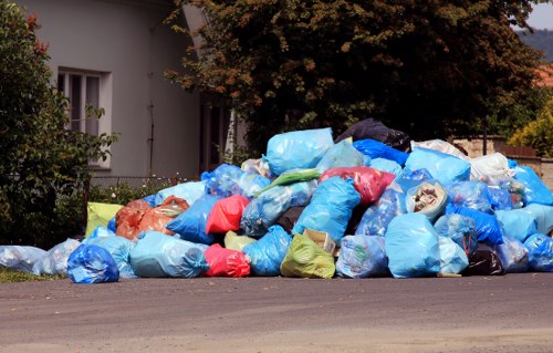 A large pile of rubbish bags, predominantly blue with some red, green, and transparent coverings, is collected on the roadside pavement in a residential area. The bags are made of plastic and appear to be filled with household waste, with some showing signs of bulging and deformation due to the contents inside. The pile is situated adjacent to a white building with visible windows and partially obscured by nearby green shrubs and a large tree with dark foliage, typical of suburban surroundings in Kentish Town. The scene features a paved street with an asphalt surface, and the rubbish bags extend from the front of the building towards the edge of the image, indicating temporary accumulation awaiting collection. The lighting suggests an overcast day, with diffuse natural light evenly illuminating the scene, aligning with urban waste collection practices often managed by local providers such as Commercial Waste Removal Kentish Town. This visual depiction effectively illustrates the type of clutter involved in commercial and residential waste disposal in London neighborhoods.