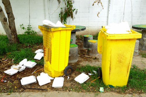 Two large, yellow plastic wheelie bins with their lids open, situated outdoors on a paved area near a white painted brick wall. The bins are used for rubbish disposal and are positioned side by side, with the left bin slightly turned to the right. Surrounding the bins, on the ground, are numerous discarded white foam takeaway containers, some open and some closed, scattered haphazardly on the concrete and grass patches. The environment shows signs of waste disposal, with ruffled grass and a few small green plants growing nearby, and a small tree trunk visible on the left. In the background, there are small, round concrete stools painted in green and yellow, and a section of green foliage climbing the wall. Overall, the scene depicts a typical outdoor rubbish collection area, emphasizing the need for professional waste management services such as those offered by Commercial Waste Removal Kentish Town to address littering and waste accumulation in the local area.