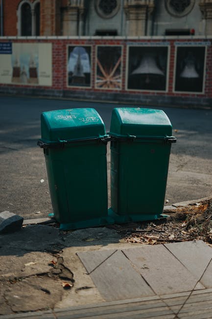 A white commercial waste collection truck with a large, open rear hopper positioned on a narrow cobblestone street in an urban area, collecting rubbish from a blue wheeled bin held by a worker wearing